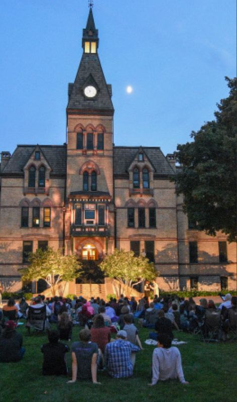 Old Main stands backdrop as a night of theater unfolds on the lawn. Students watch the performances while the light of the rising moon glows behind.